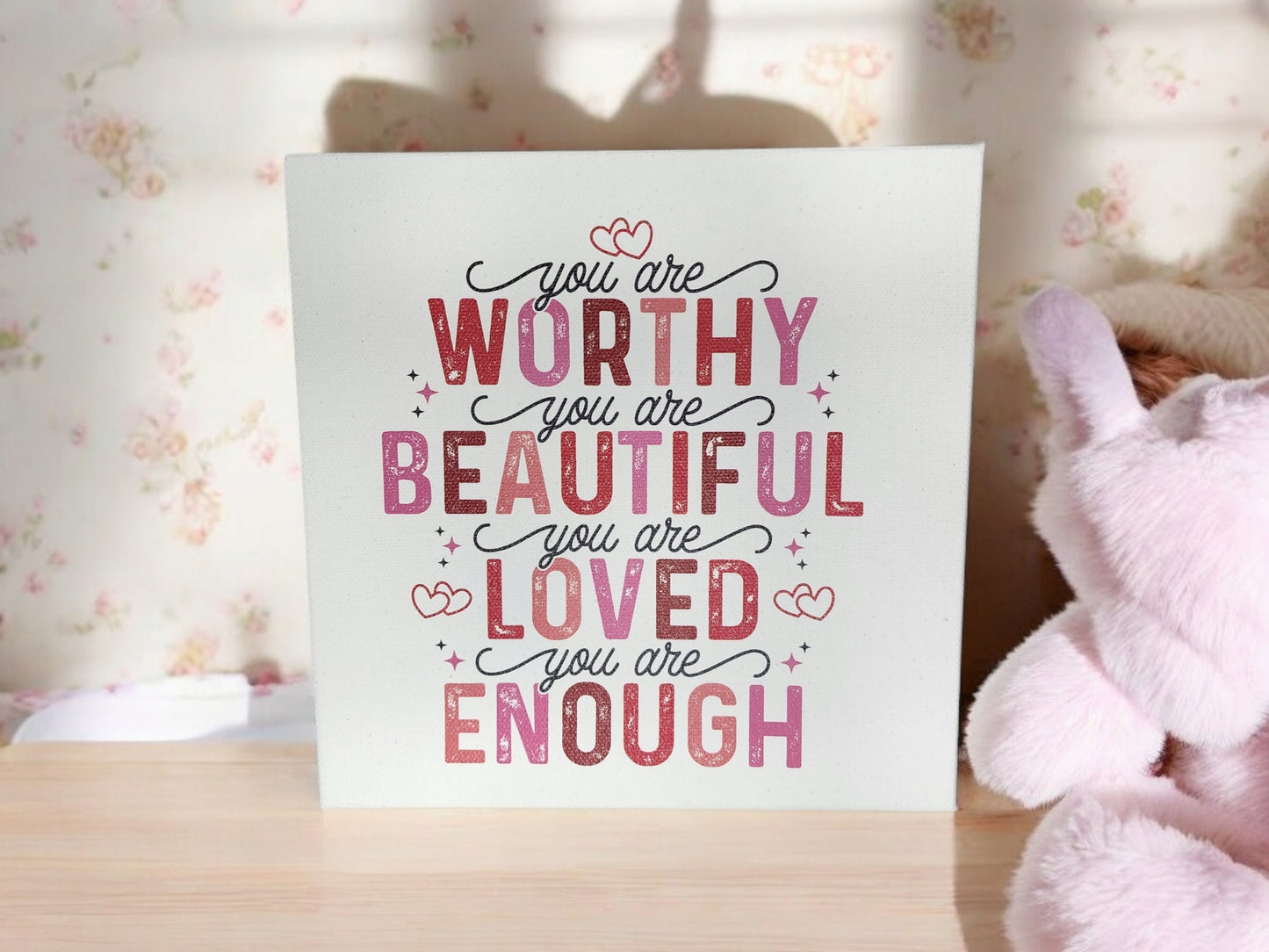 The Mirabilia Boutique 8x8 cradled canvas sign titled "You Are Worthy, Beautiful, Loved, Enough" features an empowering affirmation in pink, red, and black script. This uplifting art piece is elegantly framed by a plush toy and floral wallpaper, enhancing your home's warmth.