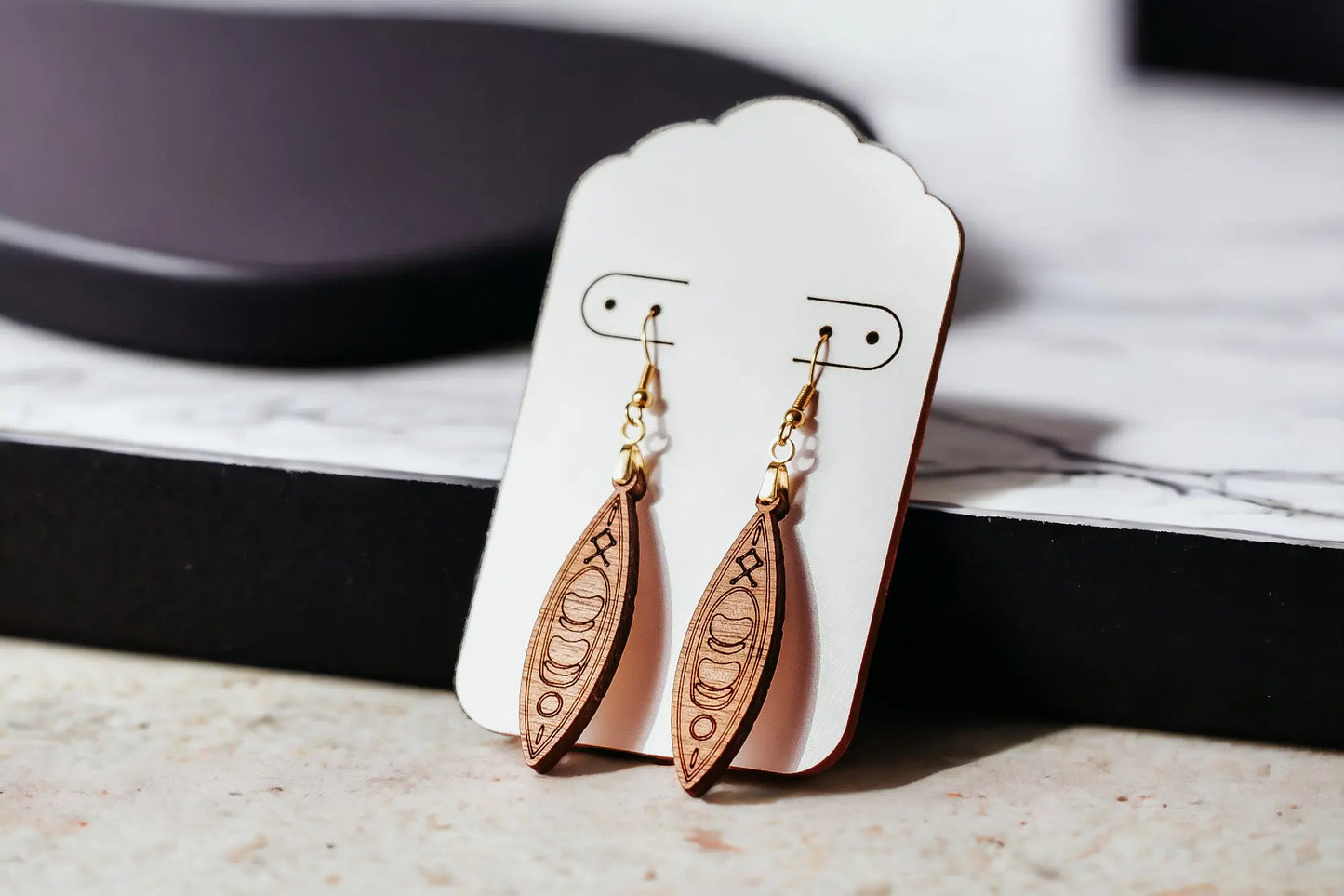 The Kayak Earrings from Mirabilia Boutique highlight exquisite moon phase designs carved into handcrafted wood. They are presented on a white display card with hypoallergenic hooks, elegantly resting on a marble surface. A blurred black tray provides a subtle backdrop enhancement.