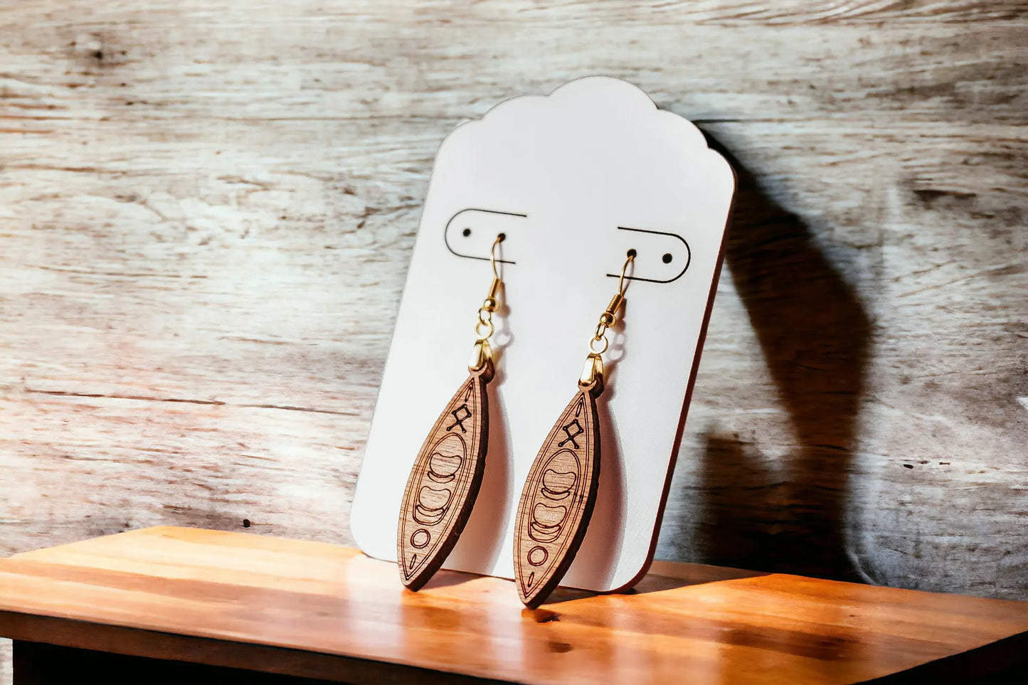 A pair of Kayak Earrings by Mirabilia Boutique, featuring intricate carvings, hang from a white display card. Handcrafted from wood, they rest on a wooden surface against a rustic background, with warm light accentuating their natural texture. Hypoallergenic hooks provide comfort for sensitive ears.