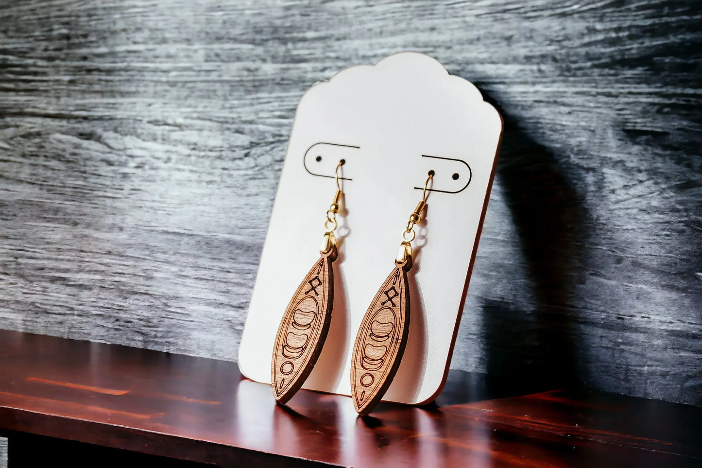 A pair of Kayak Earrings from Mirabilia Boutique, intricately carved in brown wood with crescent moon designs, is displayed on a white card with rounded edges. These earrings, featuring hypoallergenic hooks for enhanced comfort, are set against a textured gray background and rest on a dark wooden surface.