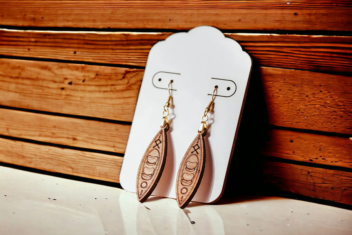 A pair of intricately carved Kayak Earrings from Mirabilia Boutique hangs on a white display card. These handcrafted wood pieces feature elongated oval shapes with detailed engravings, come with hypoallergenic hooks, and are set against a wooden background with horizontal planks.