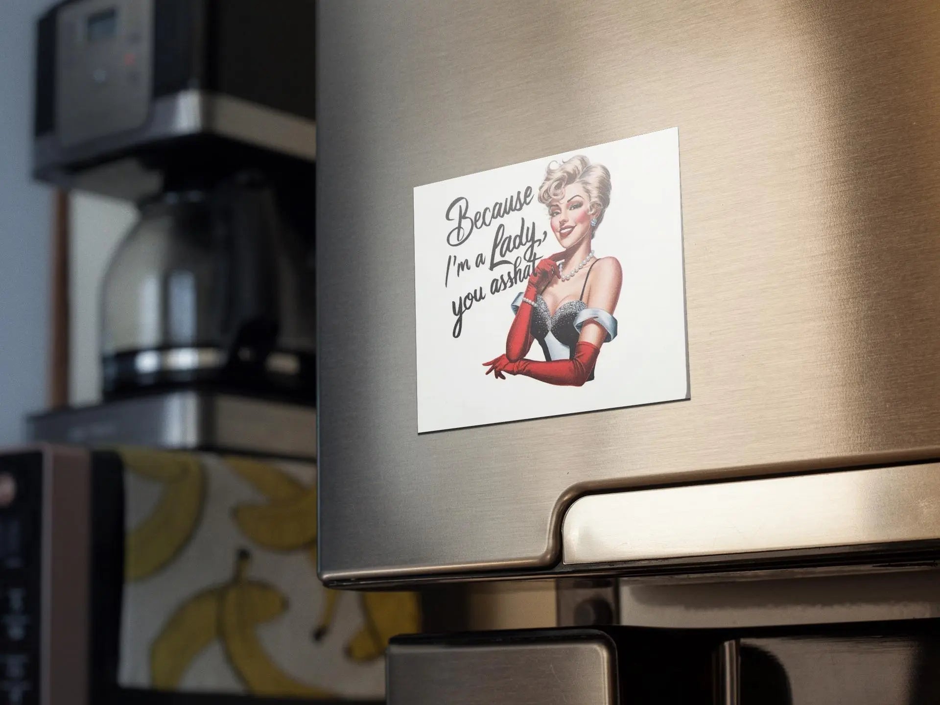 A refrigerator features the "Snarky Magnet: Because I'm a Lady You Asshat" by Mirabilia Boutique, showcasing an illustration of a vintage, glamorous woman in a red dress and gloves. The caption on the magnet humorously declares, "Because I'm a lady, you asshole!" In the background, you can spot a coffee maker and banana-themed wallpaper. It's certainly a statement piece of empowerment humor.