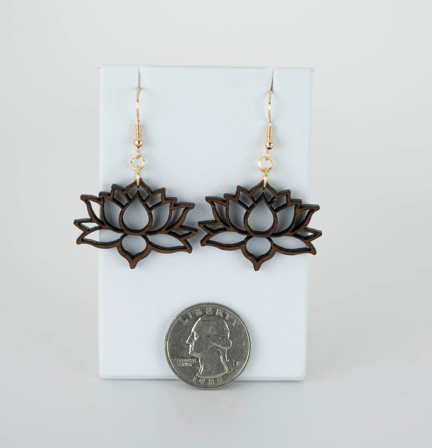 The Lotus Flower Drop Earrings by Mirabilia Boutique, featuring a wooden design and gold hooks, are elegantly showcased on a white stand. A US quarter is placed beneath them to provide a size comparison, emphasizing their delicate craftsmanship and hypoallergenic nature.