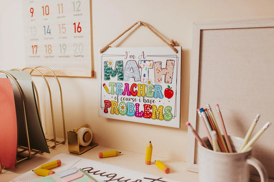 A neatly organized desk features the "I'm a Math Teacher: Humorous Problems Hanging Sign" by Mirabilia Boutique, playfully stating, "I'm a math teacher of course I have problems." The sign is surrounded by colorful pencils, papers, and a calendar, with a pencil holder and paper organizer adding to the tidy mathematical decor.