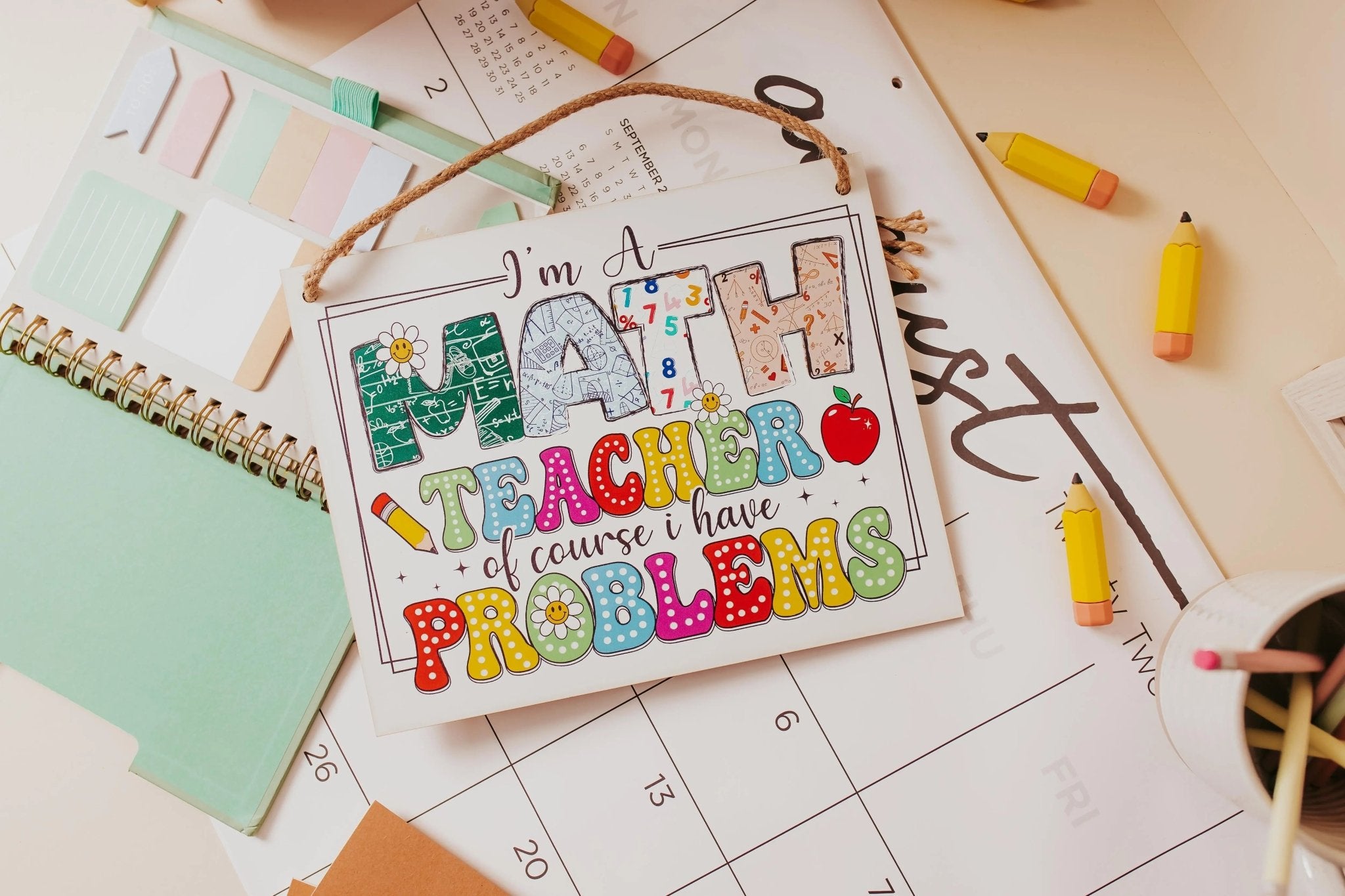 A whimsical decor piece from Mirabilia Boutique, the "I'm a Math Teacher: Humorous Problems Hanging Sign," displays the colorful phrase atop an open planner, surrounded by pencils, sticky notes, and a notebook.