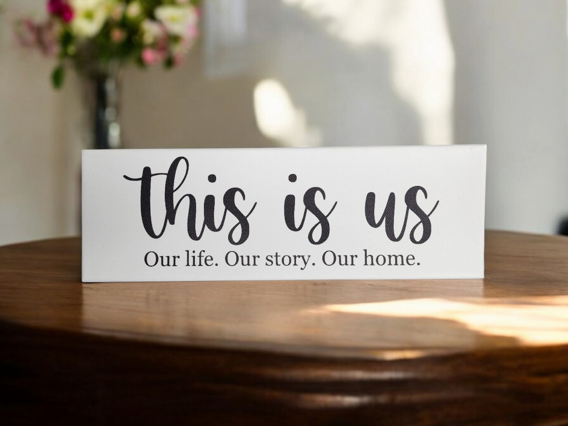 The 4x12 cradled canvas from Mirabilia Boutique, reading "this is us - Our life. Our story. Our home," is a timeless decor piece. Displayed on a wooden table, it creates a cozy atmosphere with soft shadows and a blurred floral arrangement as its backdrop.