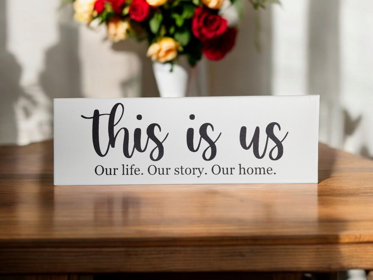 A "This Is Us. Our Life. Our Story. Our Home." 4x12 Cradled Canvas by Mirabilia Boutique sits on the table, with a vase of red, yellow, and white flowers softly blurred in the background—a perfect piece of meaningful decor that enhances timeless design.