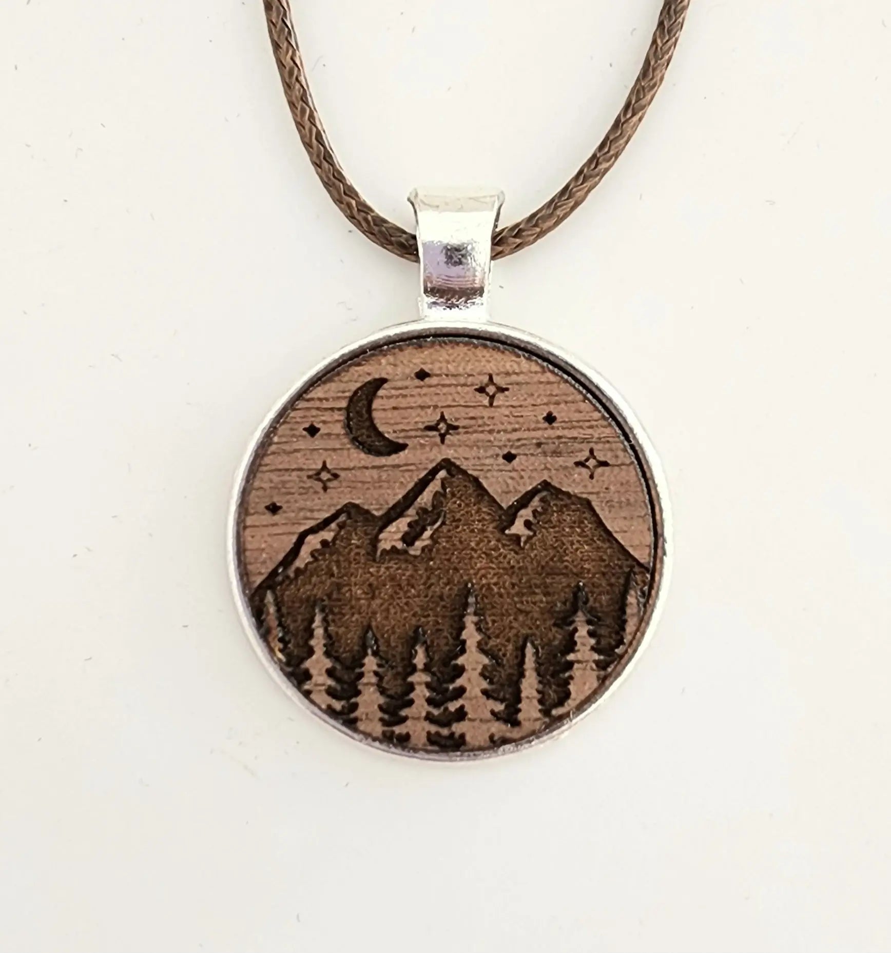 The Mountain Moon Celestial Pendant with Stars and Nature Motif from Mirabilia Boutique features an engraved image of mountain peaks, pine trees, a crescent moon, and stars on a round wooden piece suspended by a brown cord. The design is etched in a darker shade on a lighter background, creating a rustic and nature-themed look that's perfect for jewelry lovers.