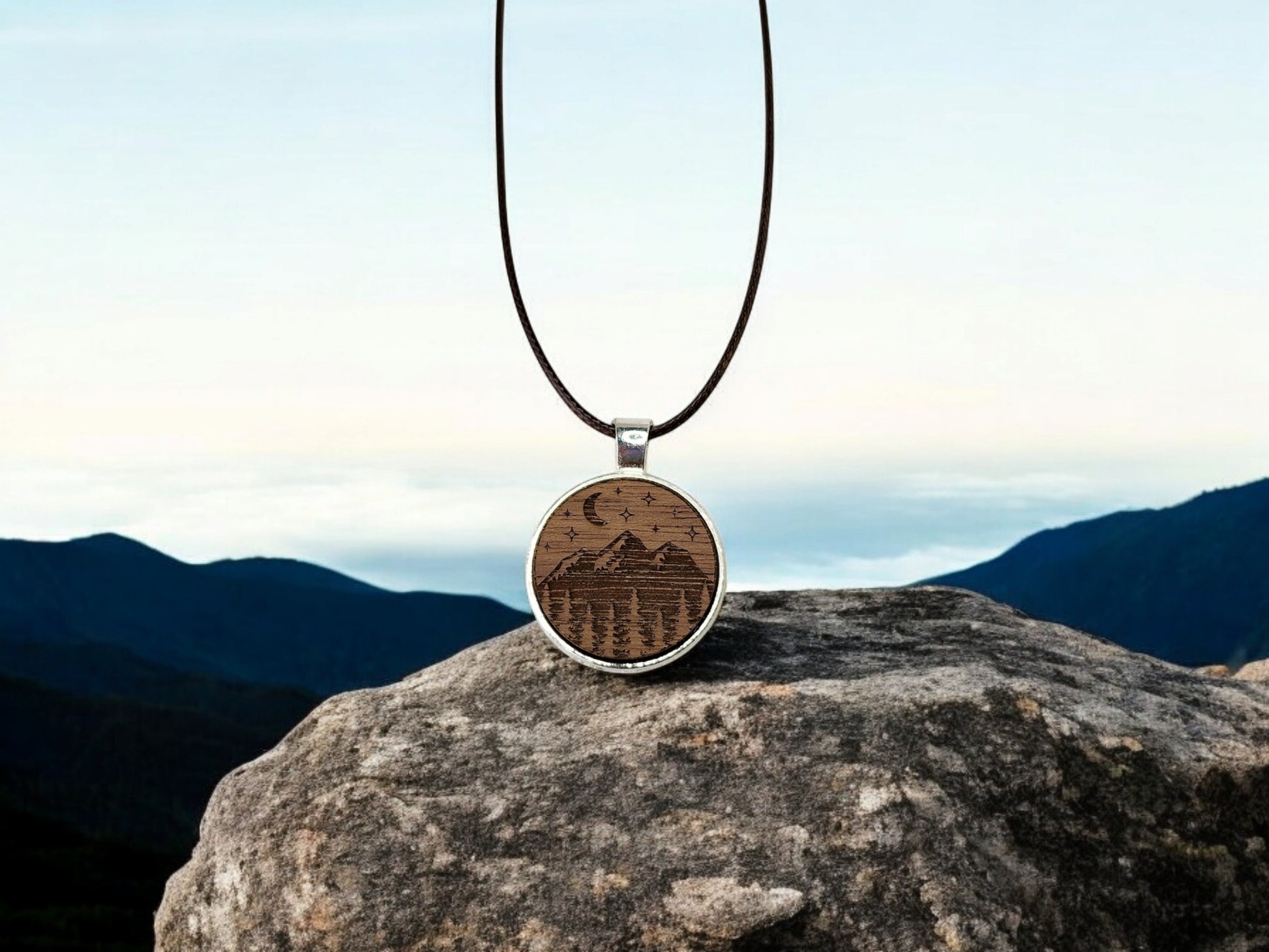 The Walnut Wood Mountain Pendant Necklace by Mirabilia Boutique features a round walnut pendant with carved mountains and trees, suspended on a leather cord. Resting on a large rock against a clear sky, it embodies the essence of nature-inspired jewelry.