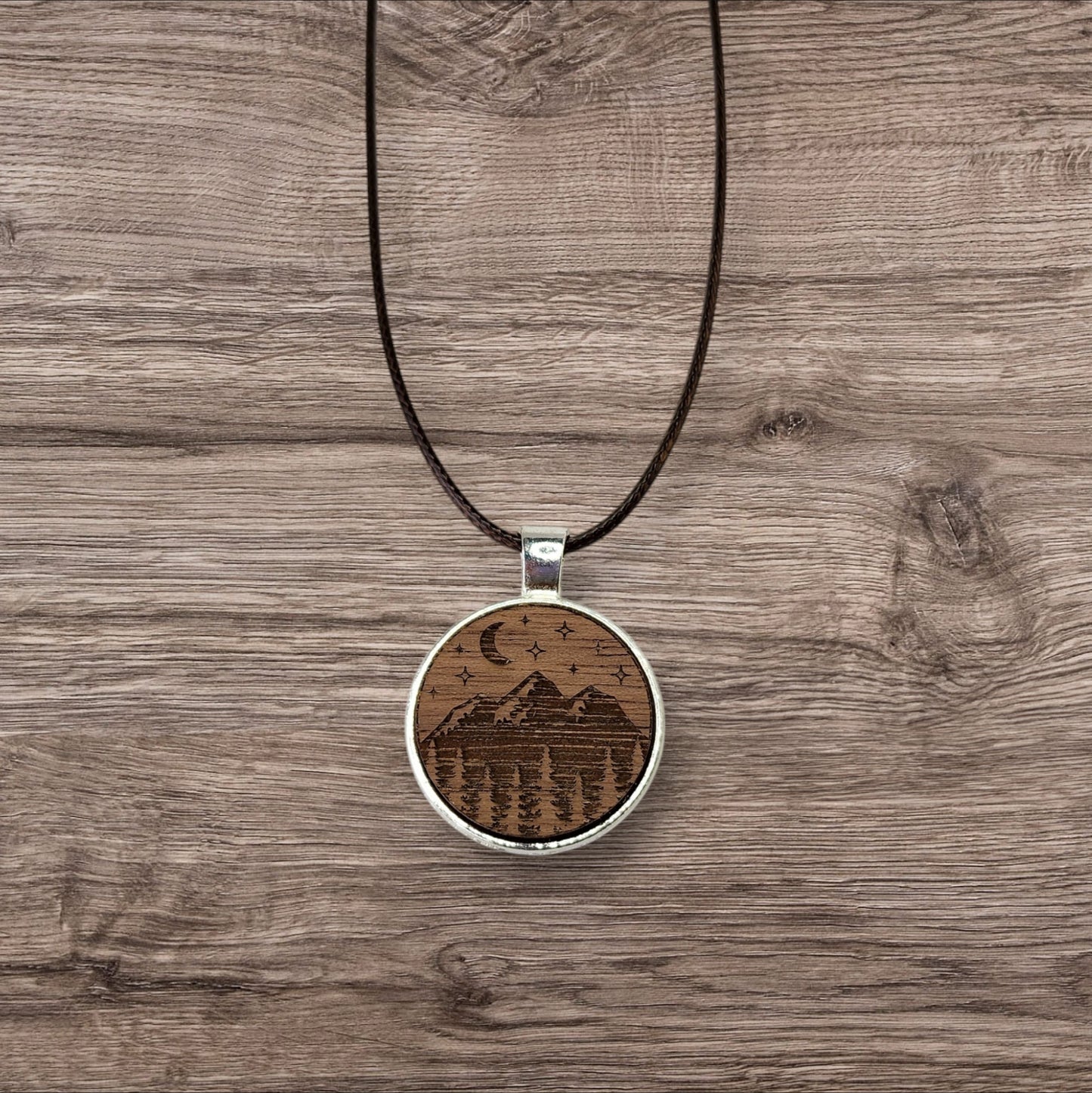 Discover the charm of nature with Mirabilia Boutique's Walnut Wood Mountain Pendant Necklace. Featuring a circular walnut pendant intricately engraved with mountains, trees, a crescent moon, and stars on a natural wooden background.