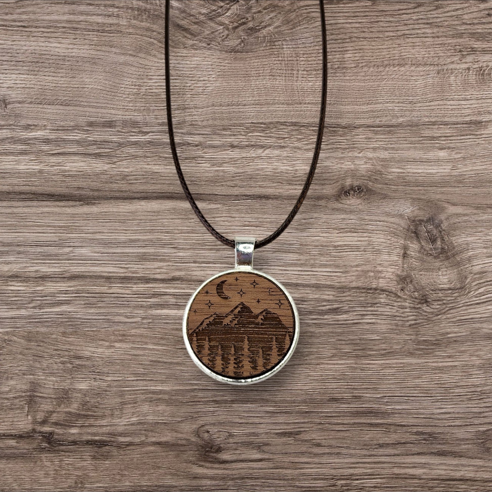 Discover the charm of nature with Mirabilia Boutique's Walnut Wood Mountain Pendant Necklace. Featuring a circular walnut pendant intricately engraved with mountains, trees, a crescent moon, and stars on a natural wooden background.