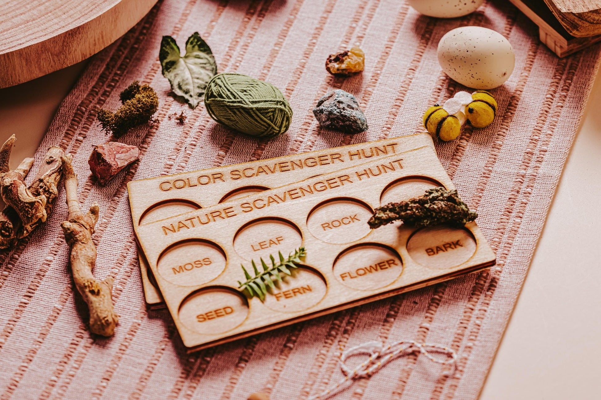 A close-up of the Kids Scavenger Hunt Bundle from Mirabilia Boutique. The wooden board features circular slots labeled "Moss," "Leaf," "Rock," "Seed," "Fern," "Flower," and "Bark." It is surrounded by various items such as yarn, eggs, leaves, and rocks on a woven fabric, making it perfect for outdoor activity games.