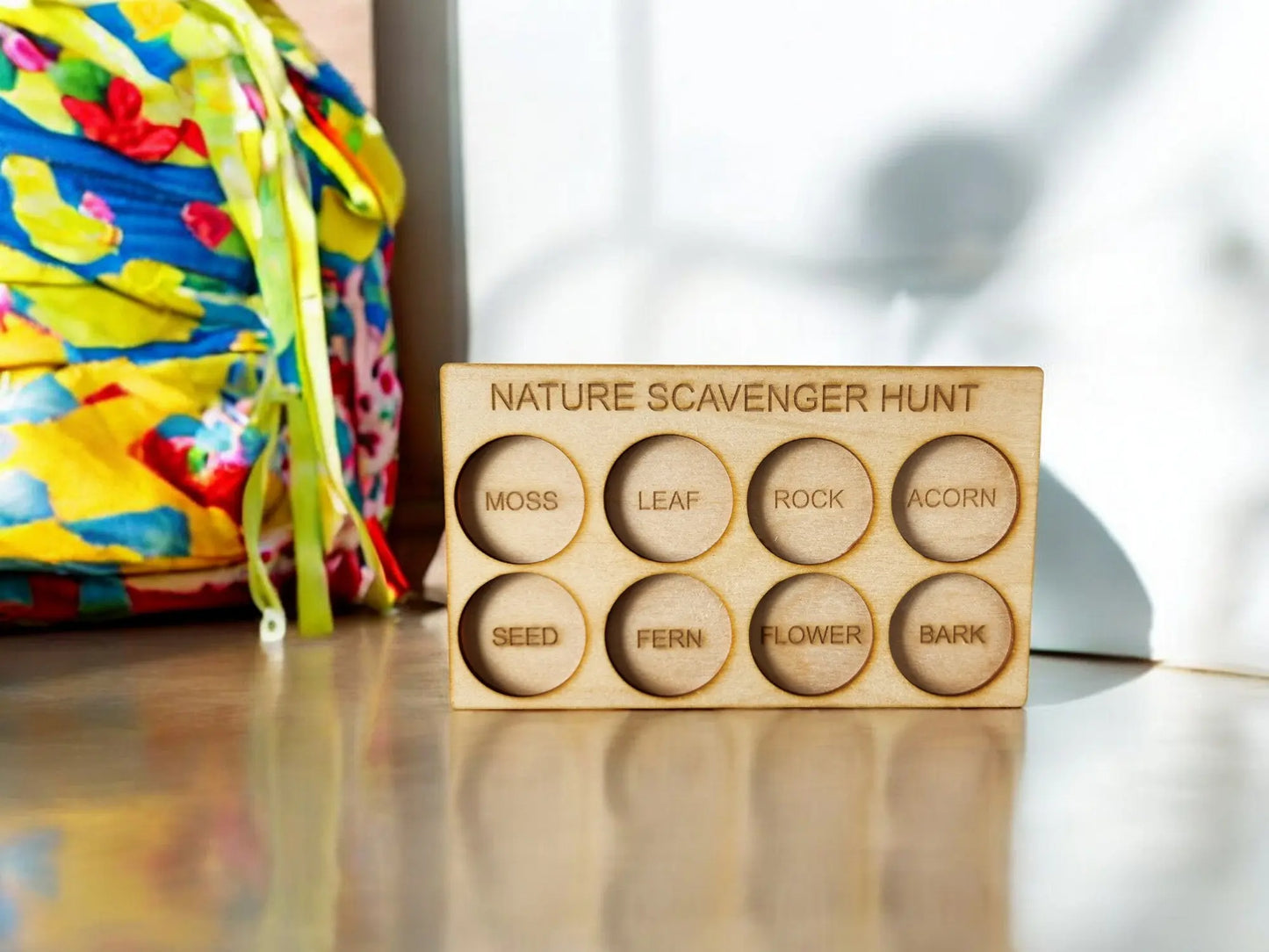 The Nature Scavenger Hunt for Kids: Fun Outdoor Adventure Game, crafted from eco-friendly wood by Mirabilia Boutique, showcases items such as moss, leaf, and rock. Set against a light wooden surface with a vibrant bag in the background, this charming outdoor toy is bathed in sunlight.