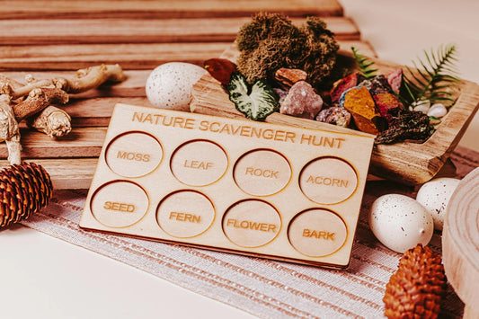 The Nature Scavenger Hunt for Kids: Fun Outdoor Adventure Game by Mirabilia Boutique features an eco-friendly wooden board with circular slots labeled "Moss," "Leaf," "Rock," and more. Surrounded by natural items like mushrooms and pinecones on a striped cloth, it makes the perfect engaging outdoor toy for curious minds eager to explore the wonders of nature.