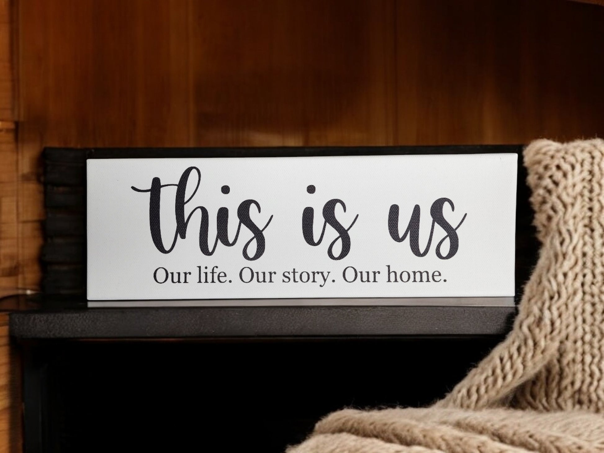 The Mirabilia Boutique's "This Is Us. Our Life. Our Story. Our Home." 4x12 cradled canvas, featuring large cursive and smaller print, is a meaningful decor piece that complements a shelf by a knitted blanket.