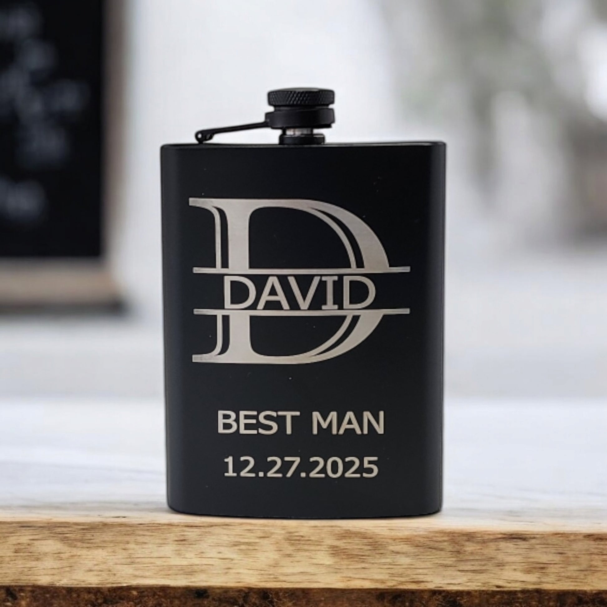 A Personalized Matte Black Hip Flask by Mirabilia Boutique, engraved with "David," "Best Man," and "12.27.2025," stands upright on a wooden surface.