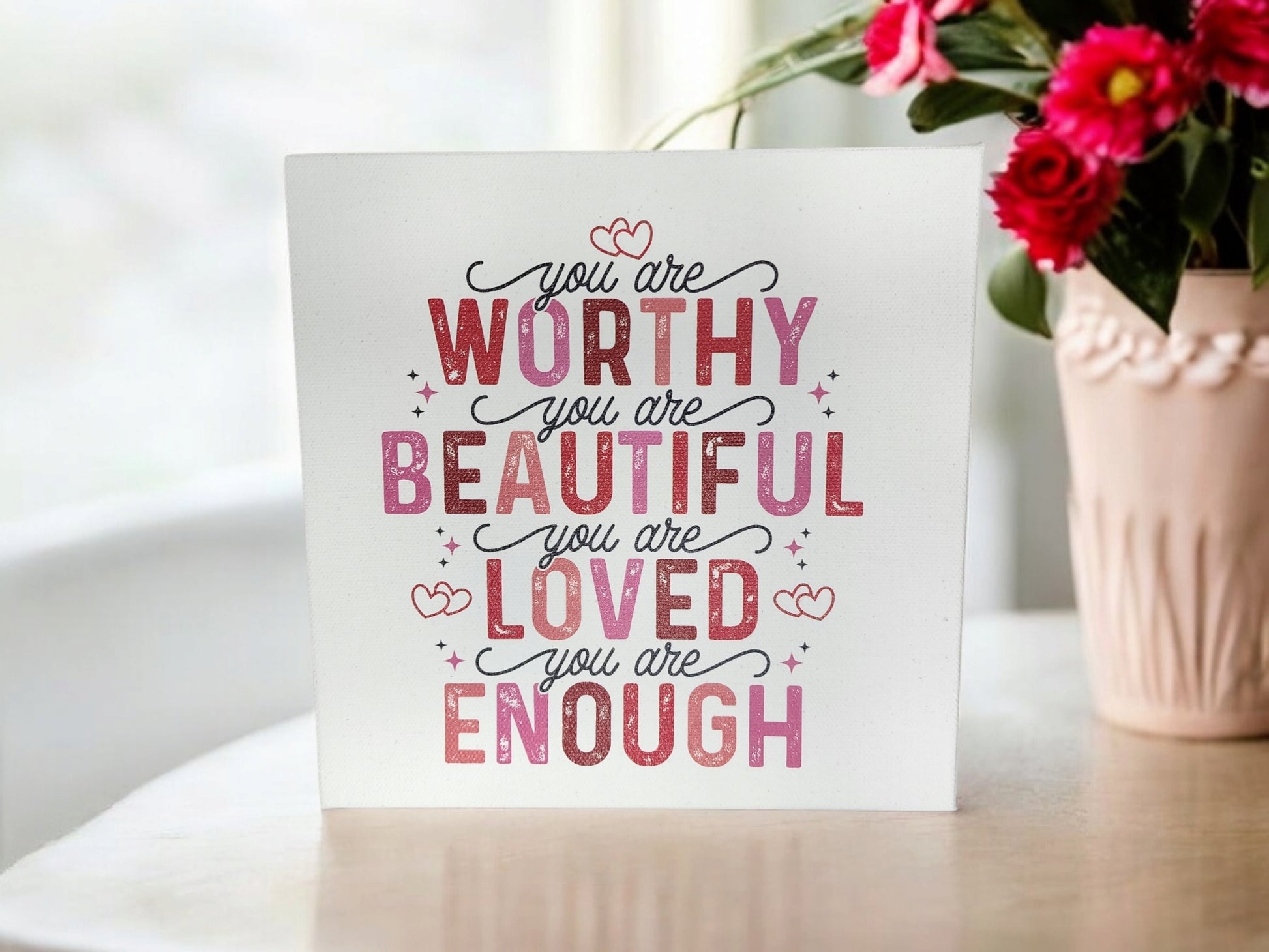 The Mirabilia Boutique's "You Are Worthy, Beautiful, Loved, Enough" 8x8 cradled canvas sign features a pink and red affirmation. Paired with a vase of pink flowers on the right, this uplifting piece enlivens any home decor with its heartfelt message.