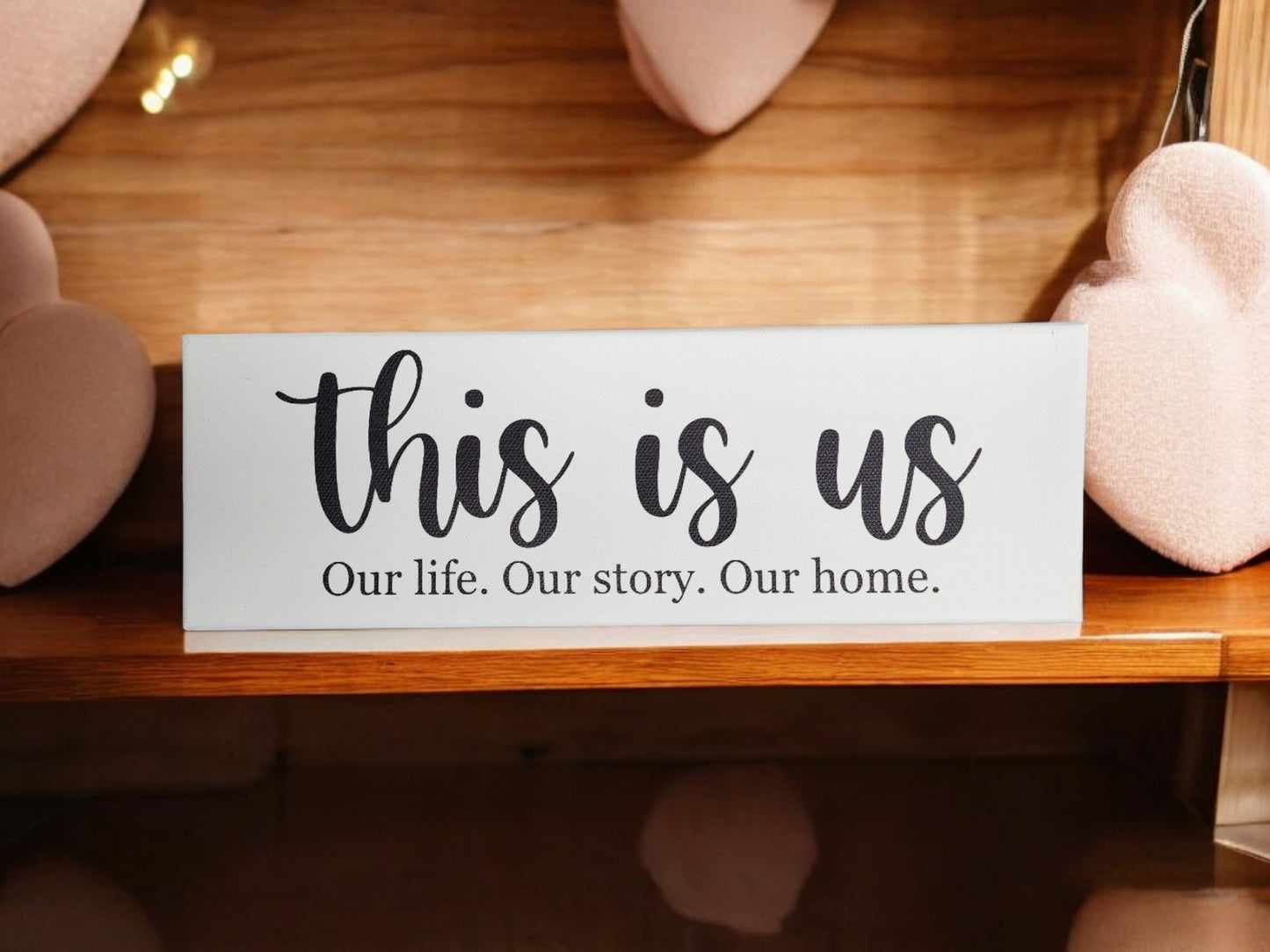 Mirabilia Boutique's "This Is Us. Our Life. Our Story. Our Home." 4x12 cradled canvas sits on a wooden shelf amid pink heart cushions, capturing the essence of meaningful decor.