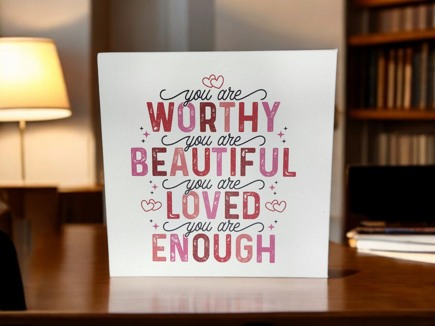 The 8x8 cradled canvas sign by Mirabilia Boutique features empowering affirmations in pink and red: "You are worthy, beautiful, loved, enough." Ideal for home decor, it sits on a wooden desk in a warmly lit room with a lamp and bookshelves.