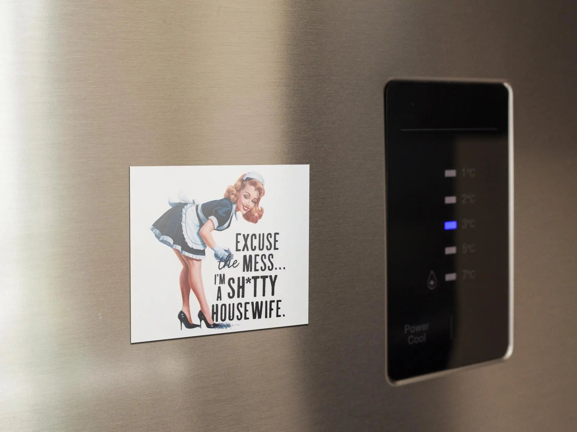 The Retro "Excuse the Mess" Shitty Housewife Magnet from Mirabilia Boutique showcases a playful woman in a maid outfit bowing, paired with the cheeky message, "Excuse the mess... I'm a sh*tty housewife." This humorous fridge decor serves as a charming and quirky kitchen accessory perfect for any stainless steel refrigerator.