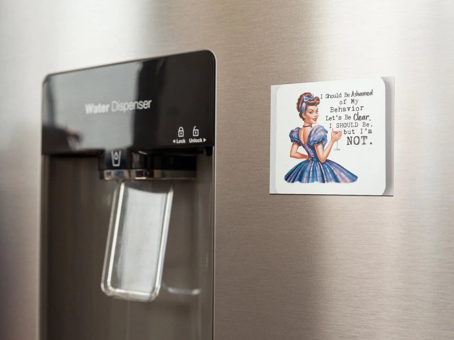 A water dispenser is mounted on a stainless steel refrigerator door. Beside it, a snarky retro magnet from Mirabilia Boutique depicts a woman in a vintage dress with humorous text: "I Should Be Ashamed of My Behavior But I'm Not.