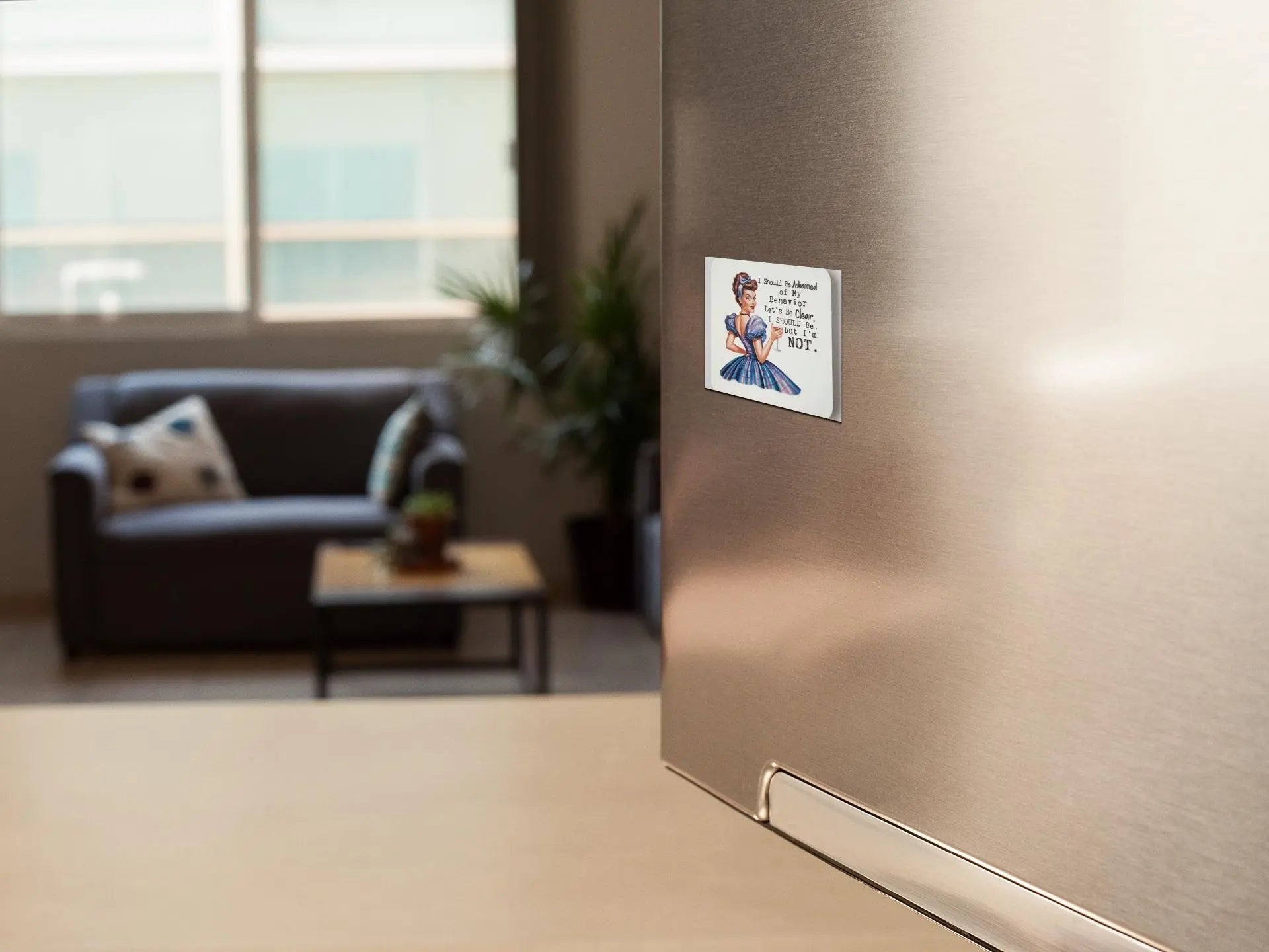 A modern kitchen scene with a stainless steel refrigerator in the foreground, adorned with Mirabilia Boutique's snarky retro magnet that reads "I Should Be Ashamed of My Behavior But I'm Not." The background features a living room area with a dark sofa, coffee table, plants, and large window. The space appears bright and airy, effortlessly combining retro flair with contemporary design.