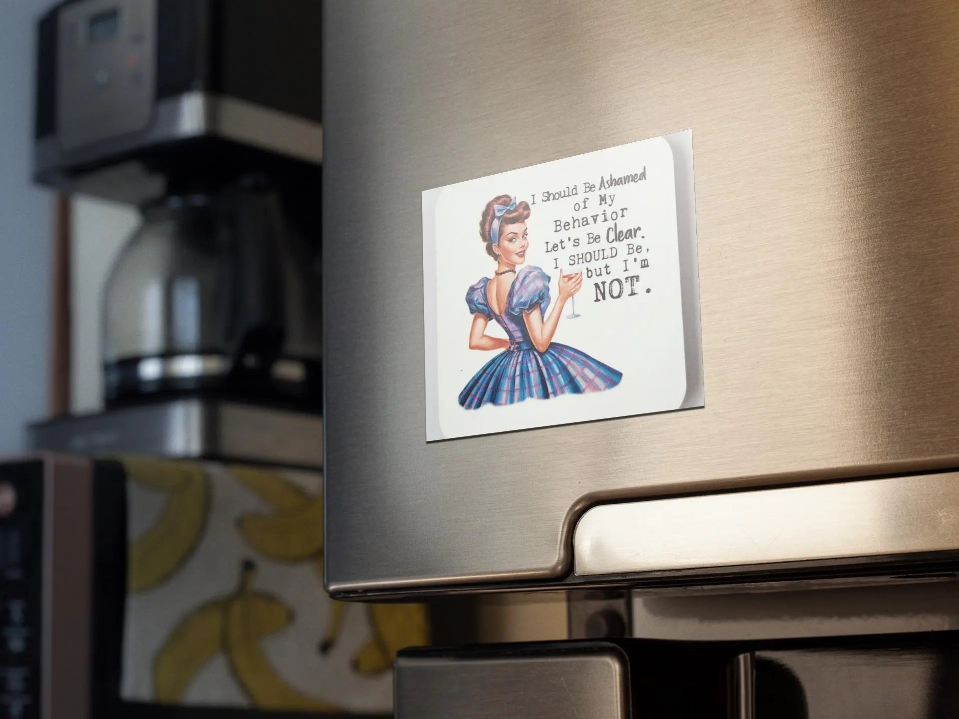 A refrigerator door adorned with a snarky retro magnet from Mirabilia Boutique, featuring a woman in a blue dress. The magnet's text boldly declares, "I Should Be Ashamed of My Behavior But I'm Not." Retro flair meets humor, while a coffee maker sits in the background.