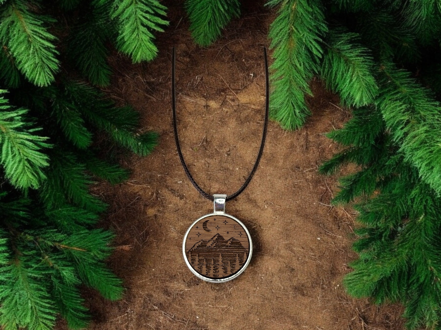 A Walnut Wood Mountain Pendant Necklace by Mirabilia Boutique rests on brown soil, encircled by green fir branches. The nature-inspired design features a mountain scene with a crescent moon and stars, hanging on a black cord to complement the rustic and natural setting.