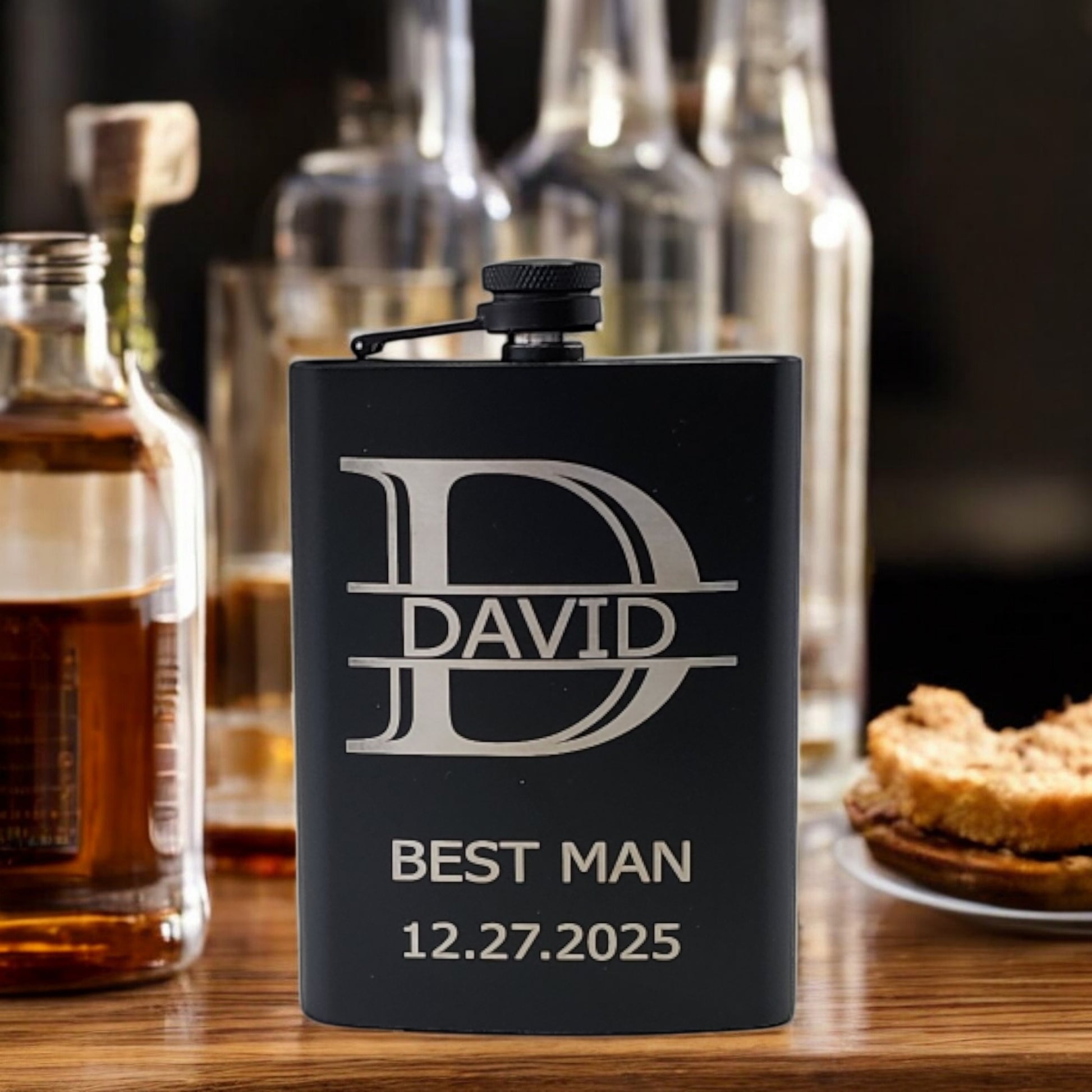 A Personalized Matte Black Hip Flask by Mirabilia Boutique, custom engraved with "David, Best Man, 12.27.2025," sits on a wooden surface surrounded by assorted glass bottles and a small plate of pie pieces, creating an inviting display.