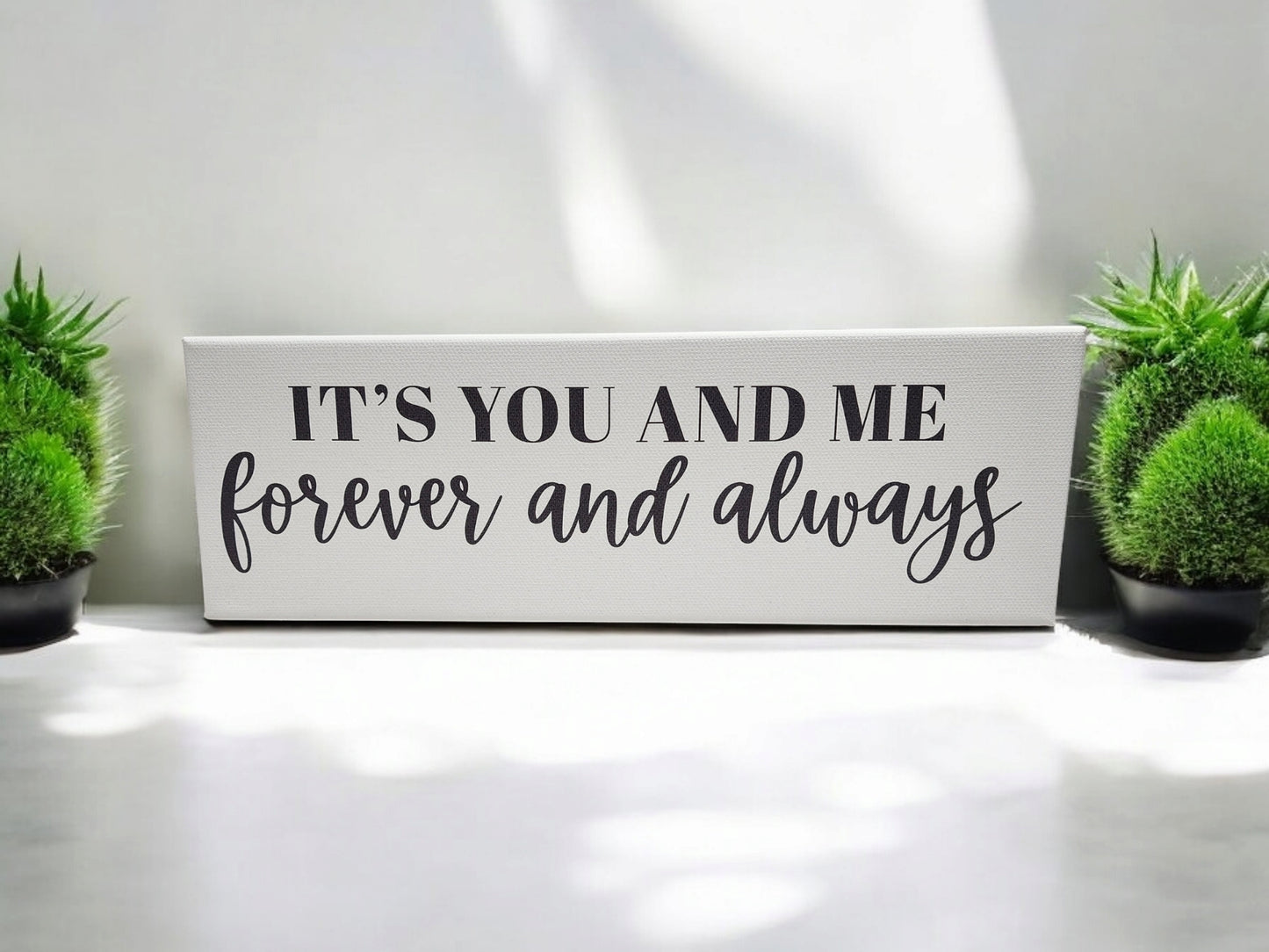 Mirabilia Boutique's "It's You and Me Forever and Always" 4x12 cradled canvas features bold, cursive black typography on a white background. The romantic wall art is styled with two small green plants that cast soft shadows, enhancing its personalized home decor appeal.