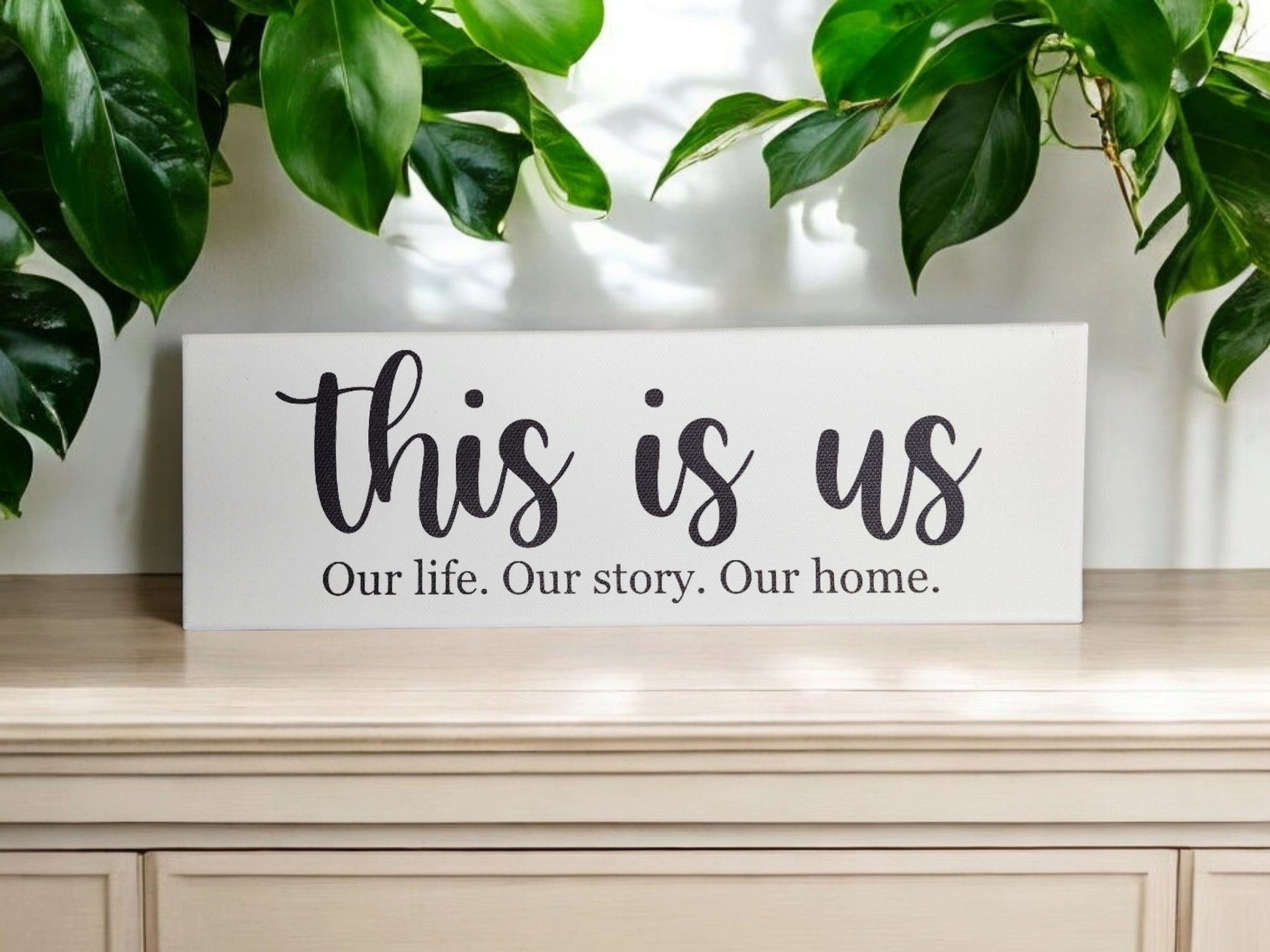 A Mirabilia Boutique 4x12 cradled canvas, "This Is Us. Our Life. Our Story. Our Home." rests on a light wooden table, framed by green leaves, showcasing timeless design and meaningful decor.