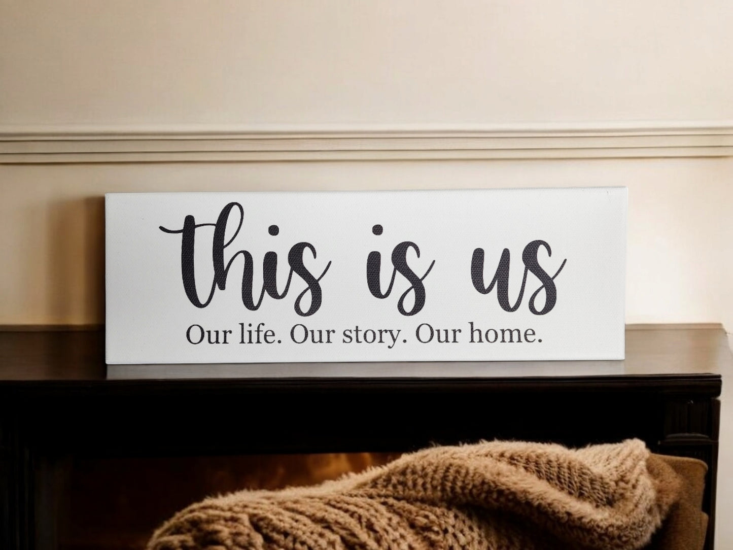 A 4x12 cradled canvas from Mirabilia Boutique, featuring "This Is Us. Our Life. Our Story. Our Home." in large script, is displayed on the mantel as meaningful decor, while a textured beige throw blanket adds warmth against a softly blurred background.