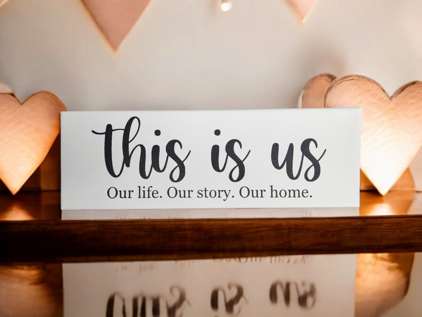 The "This Is Us. Our Life. Our Story. Our Home." 4x12 Cradled Canvas by Mirabilia Boutique rests on wood, flanked by warm-lit heart lamps, offering meaningful decor that speaks to the heart of any home with a timeless design.