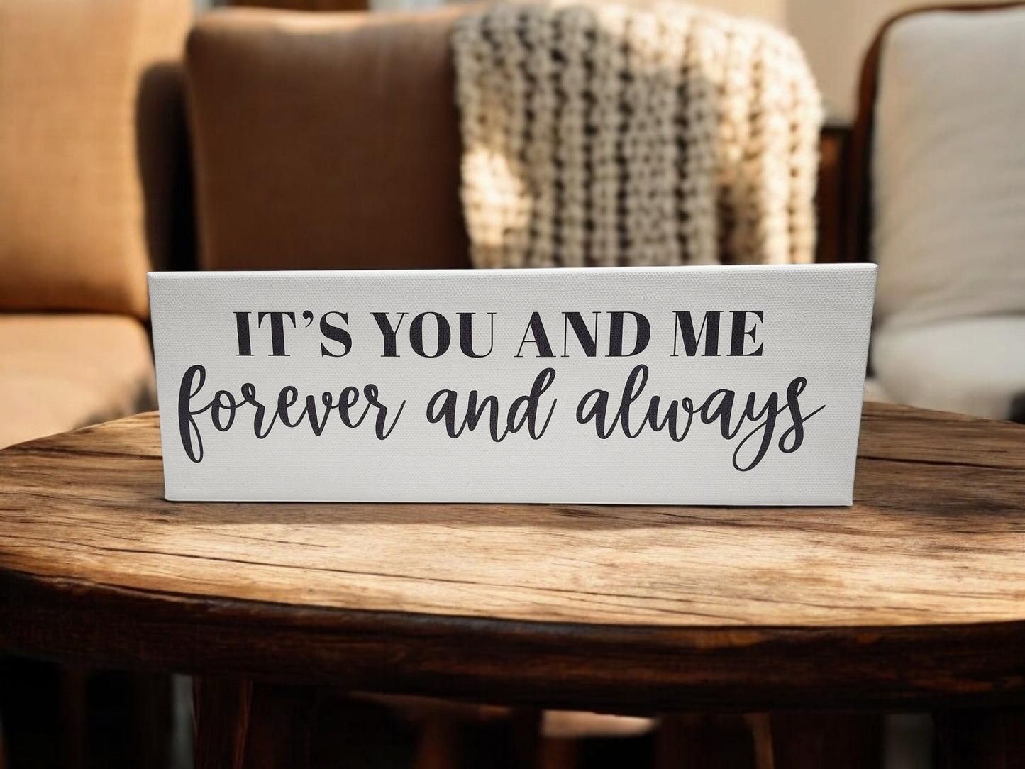 A 4x12 cradled canvas from Mirabilia Boutique, reading "It's You and Me Forever and Always," sits on a wooden table with beige chairs softly blurred in the background, adding charm to this romantic wall art.