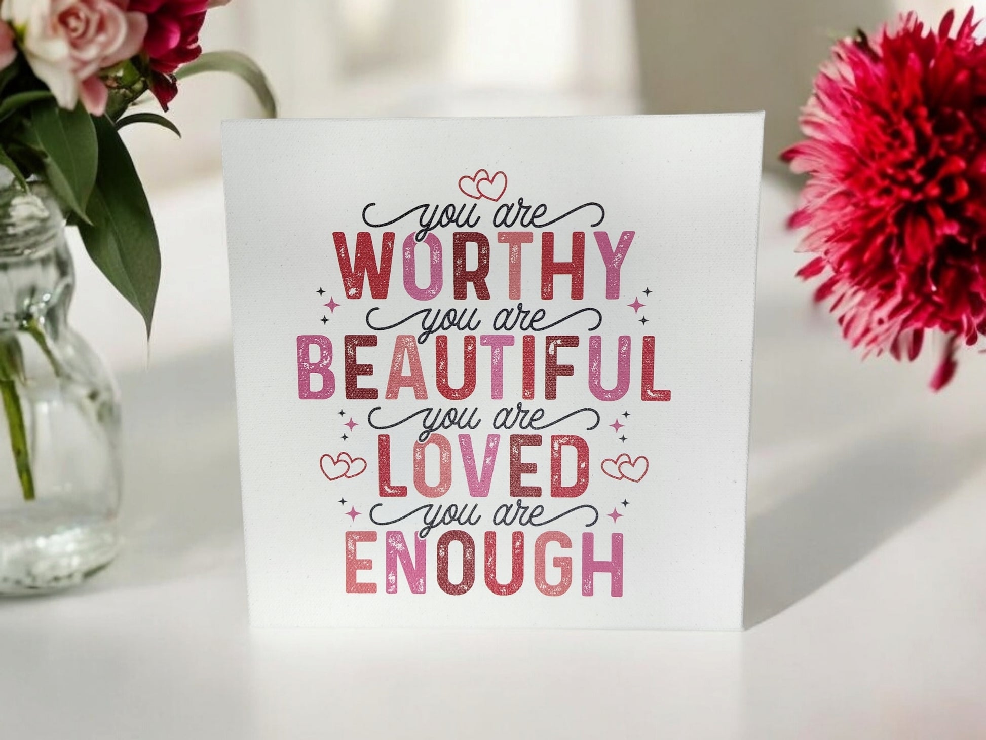 The "You Are Worthy, Beautiful, Loved, Enough" 8x8 cradled canvas sign by Mirabilia Boutique features motivational text in pink and red. Surrounded by pink flowers and a large bloom on a bright surface, it serves as both uplifting home decor and an empowering affirmation.