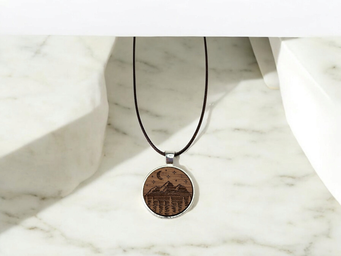 The Walnut Wood Mountain Pendant Necklace from Mirabilia Boutique features a round design with mountains, trees, and birds on a dark cord, set against marble. Its nature-inspired theme captures the wilderness essence elegantly.