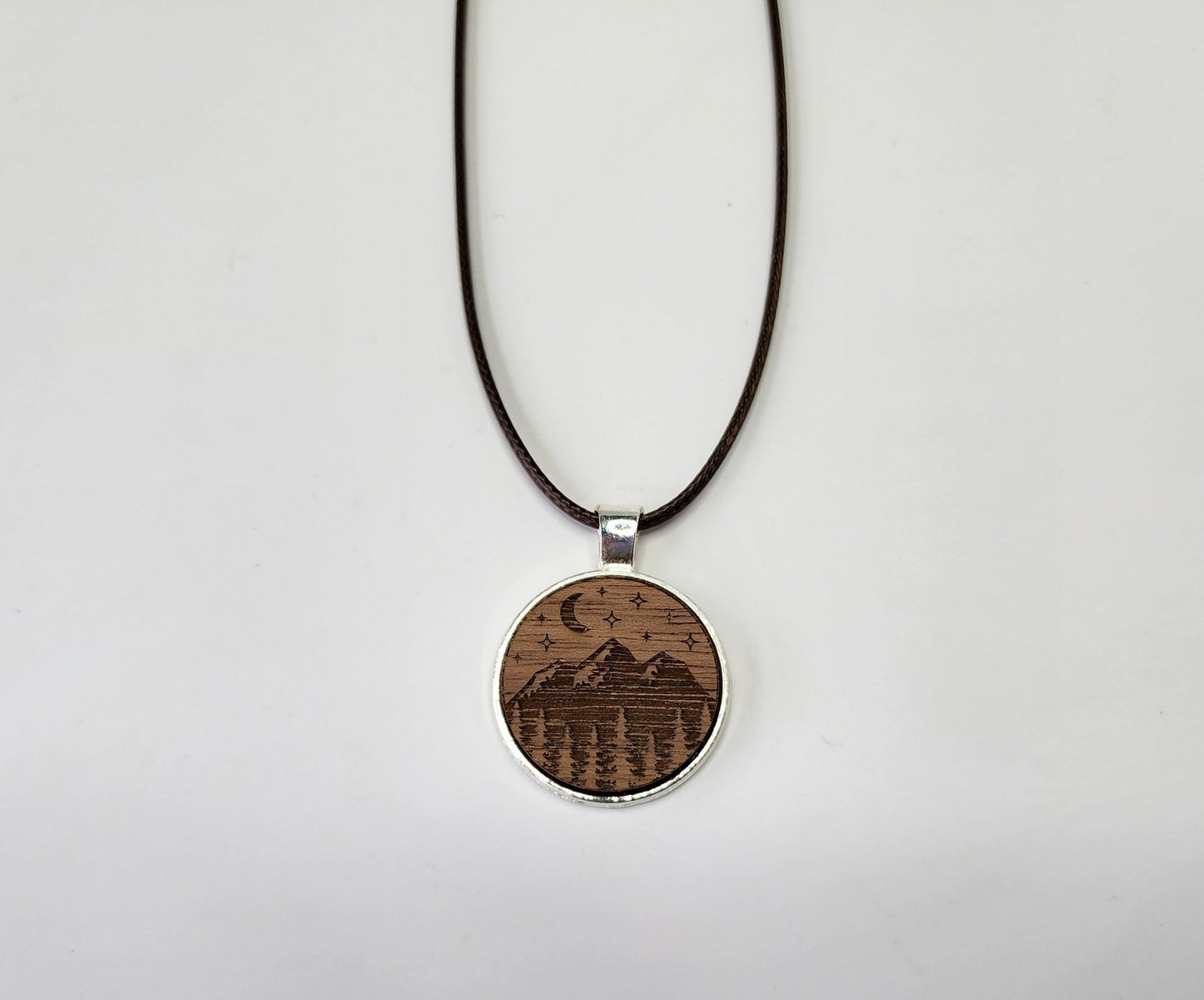 The Mirabilia Boutique Walnut Wood Mountain Pendant Necklace features a circular walnut wood pendant with an engraved mountain scene, crescent moon, stars, and trees on a black cord. The nature-inspired design stands out against a plain white background.