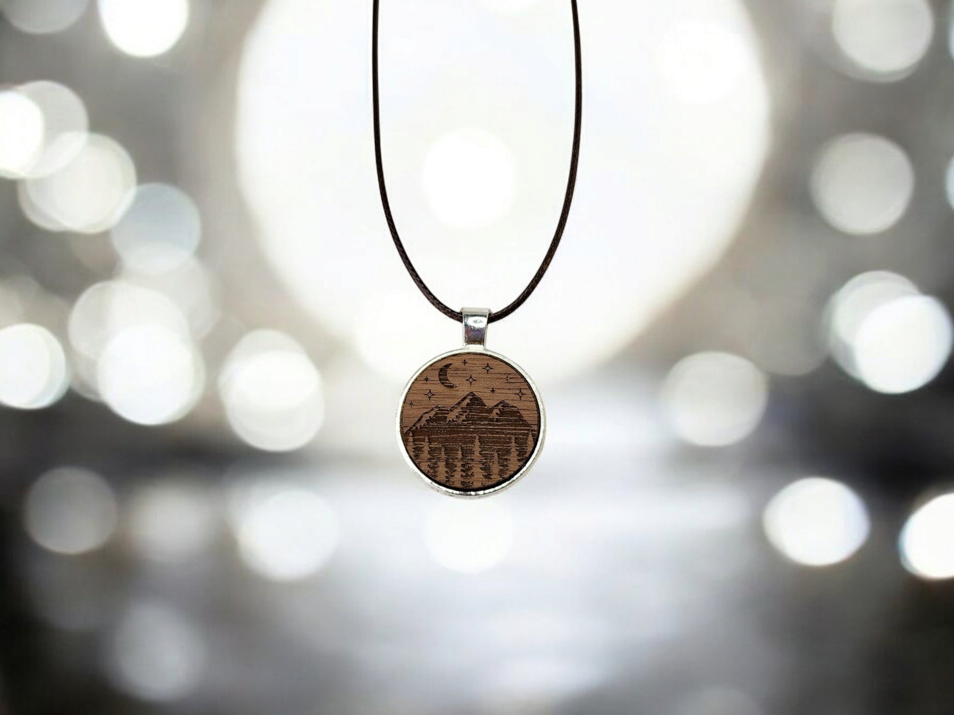 The Mirabilia Boutique's Walnut Wood Mountain Pendant Necklace, featuring a crescent moon and stars design, hangs elegantly from a dark cord with a softly blurred background of bokeh lights enhancing its serene nature-inspired appeal.