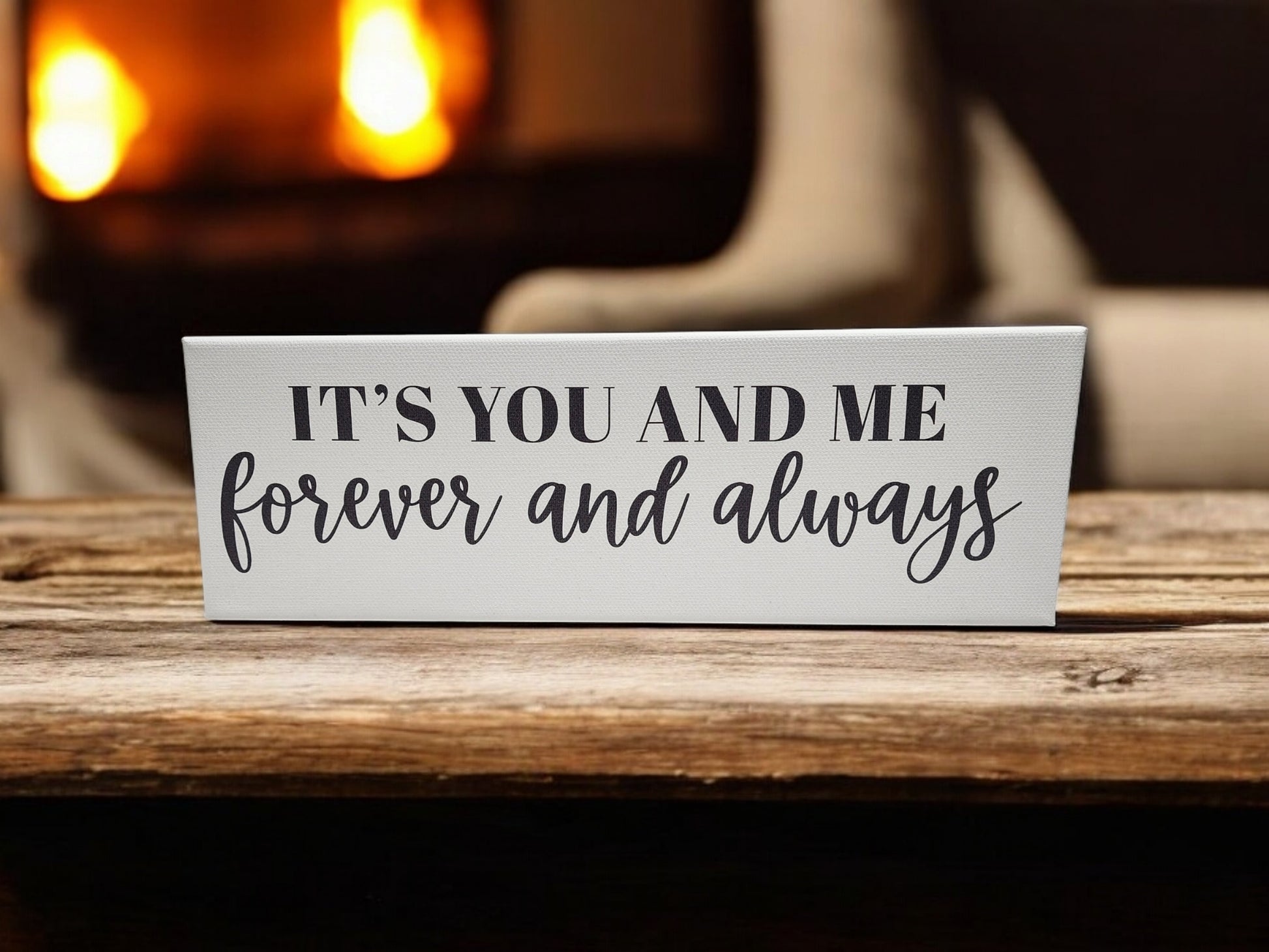 A 4x12 cradled canvas from Mirabilia Boutique, featuring the phrase "It's You and Me Forever and Always" in black on white, rests on a wooden surface. The warm, blurred background with orange light hints at a cozy ambiance, perhaps near a fireplace.