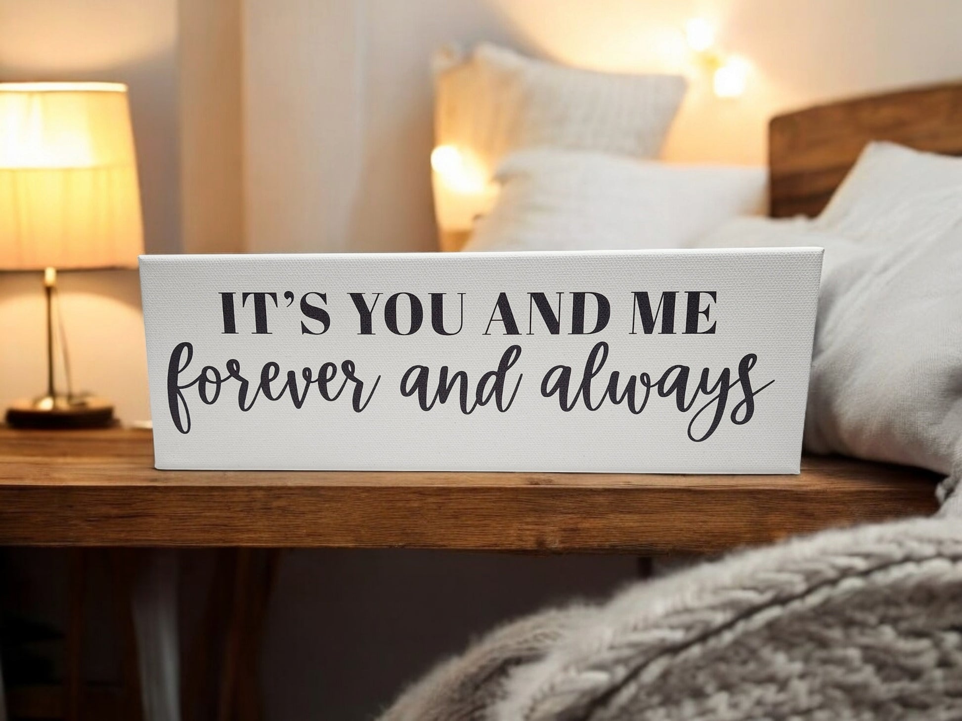 A cozy bedroom scene showcases a Mirabilia Boutique "It's You and Me Forever and Always." cradled canvas with elegant black typography. A warm lamp and neatly made bed with soft pillows and blankets complete the inviting atmosphere alongside the wooden nightstand.