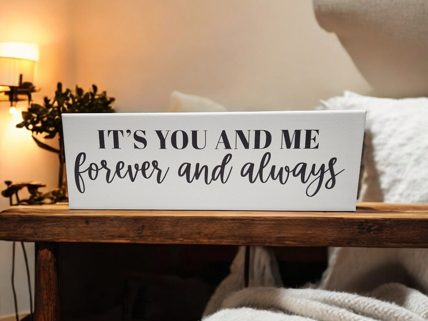 A 4x12 cradled canvas by Mirabilia Boutique reads, "It's You and Me Forever and Always." With bold black typography and script accents, it's set in a warm ambiance with soft lighting and a plant in the background, making it ideal personalized home decor.