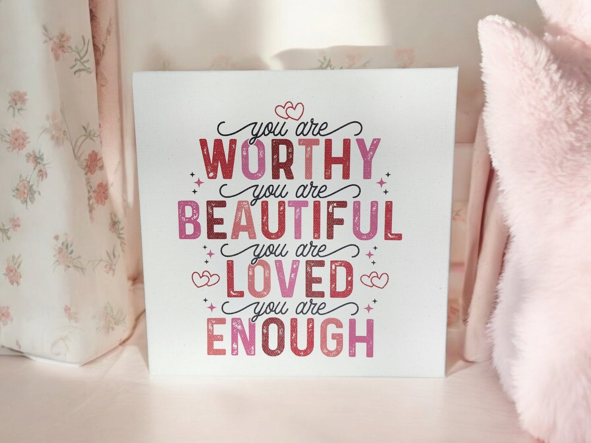 An uplifting 8x8 "You Are Worthy, Beautiful, Loved, Enough" cradled canvas sign by Mirabilia Boutique shines with pink and red lettering. It sits on a light surface near floral curtains and a fluffy pink object.